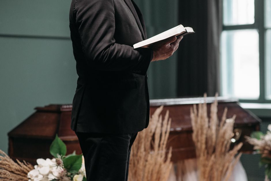 A person dressed in a black suit holding an open book or program, standing indoors near a dark wooden funeral casket adorned with floral arrangements. The flowers include white roses and green foliage