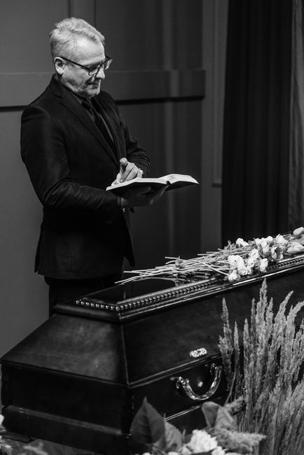 A man with grey hair and glasses, dressed in a dark suit, stands beside a closed casket during a funeral service, reading from a book held in his hands. The casket is adorned with a floral arrangement