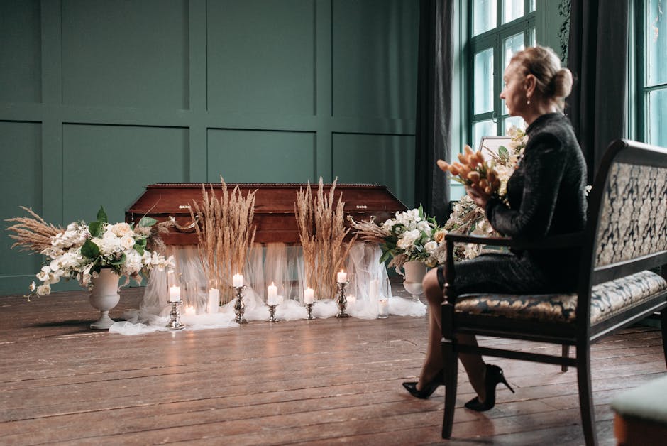 A woman seated on an ornate floral-patterned armchair holding a small bouquet of fresh flowers, with her profile visible. In front of her, there is a funeral arrangement on the floor featuring white t