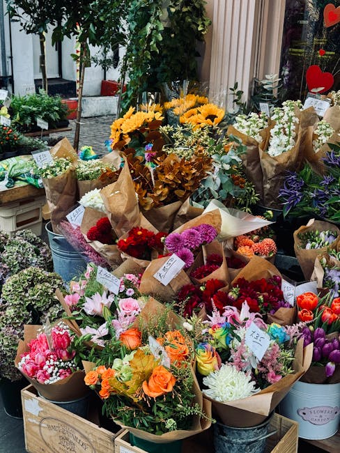 An outdoor flower display featuring a variety of fresh, colorful floral arrangements arranged in brown kraft paper wraps and white buckets, placed on wooden crates and metal tubs. The bouquets include