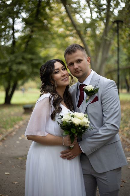 A newlywed couple standing outdoors in a park during daytime, with tall trees and greenery in the background. The bride is holding a bouquet of white roses with lush green foliage, wearing a flowing w