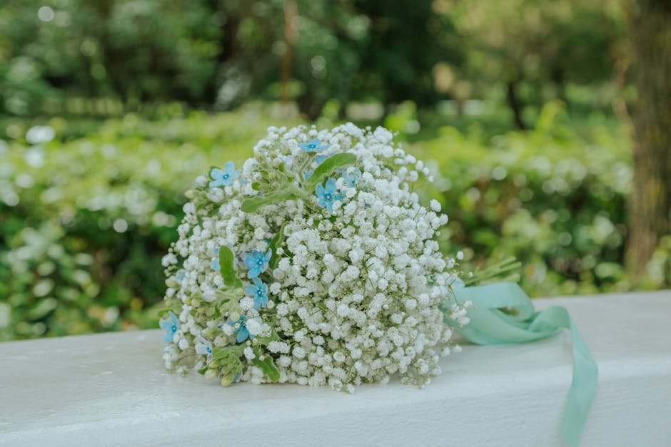A bouquet of fresh white gypsophila (baby's breath) with delicate tiny blossoms, accented with small light blue forget-me-not flowers and green foliage. The bouquet is tied with a pale blue satin ribb